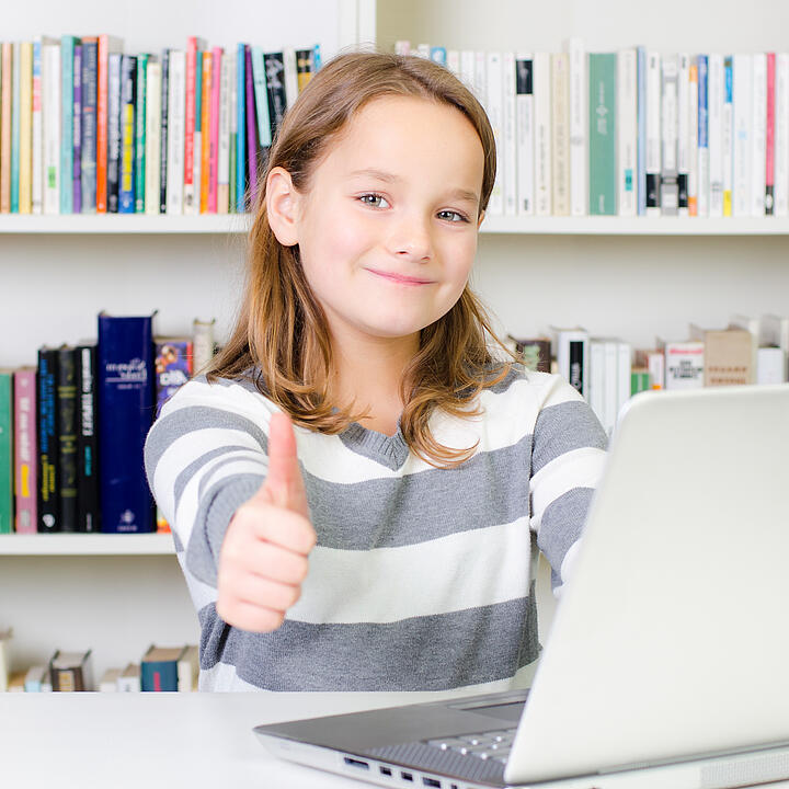 Ein junges Mädchen sitzt an einem Laptop vor einem Bücherregal und streckt den Daumen in die Kamera.